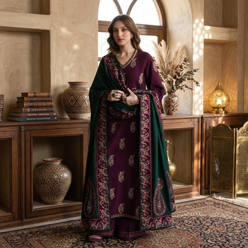 A woman stands indoors wearing a traditional outfit with a deep purple kurta featuring ornate gold and green paisley embroidery, paired with a dark green dupatta and matching embroidered border. The setting is a warm, rustic room with wooden shelves, decorative vases, and a patterned rug.