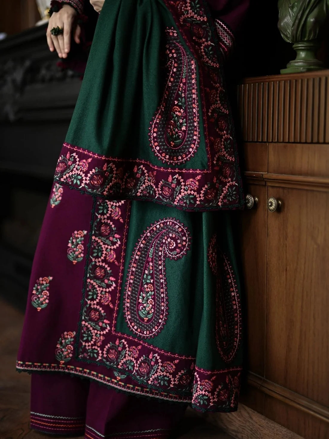 A woman stands indoors wearing a traditional outfit with a deep purple kurta featuring ornate gold and green paisley embroidery, paired with a dark green dupatta and matching embroidered border. The setting is a warm, rustic room with wooden shelves, decorative vases, and a patterned rug.